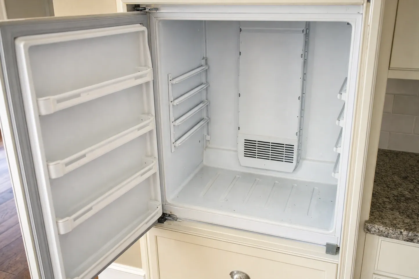 Built-in refrigerator compartment standing open, empty liner with door shelves and the evaporator vent cover behind