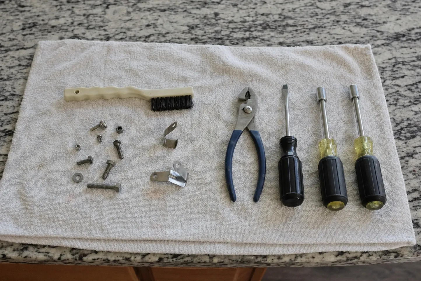 Bristle brush, slip-joint pliers, a screwdriver and two nut drivers on a towel with sorted screws and small brackets