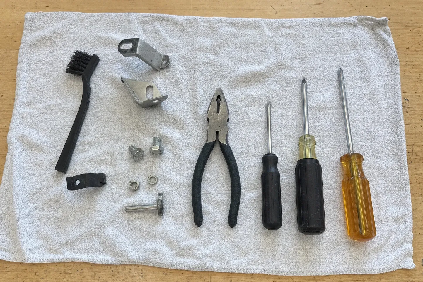 Black bristle brush, two steel brackets, bolts, nuts and washers, pliers and three screwdrivers on a folded towel