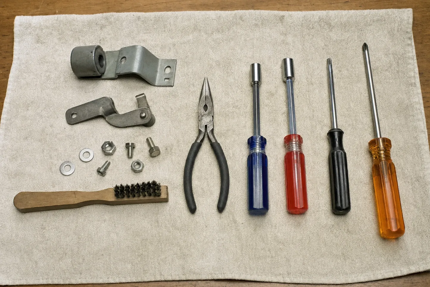 Overhead flat-lay on a cream towel of two steel hinge brackets, one carrying a roller bushing, beside needle-nose pliers, two nut drivers, a flat and a cross-head screwdriver, a wire brush and sorted bolts, nuts and washers