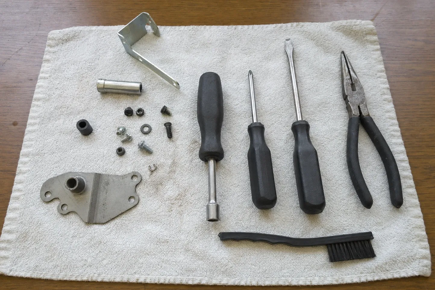 Flat-lay on a cream towel over dark wood of an angled bracket, a steel sleeve and a cast bracket with a threaded boss, with nuts, screws and a washer beside a nut driver, two screwdrivers, needle-nose pliers and a black bristle brush