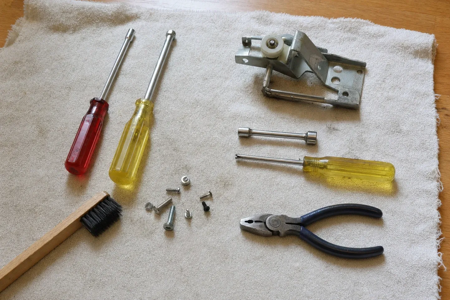 Flat-lay on a pale towel of a hinge assembly carrying a white nylon roller, with two nut drivers, a short double-ended socket, blue-handled linesman pliers, a wooden-handled brush and a scatter of small screws